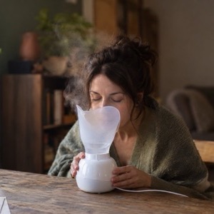 A woman leans over a small steam inhaler at a wooden table by a window